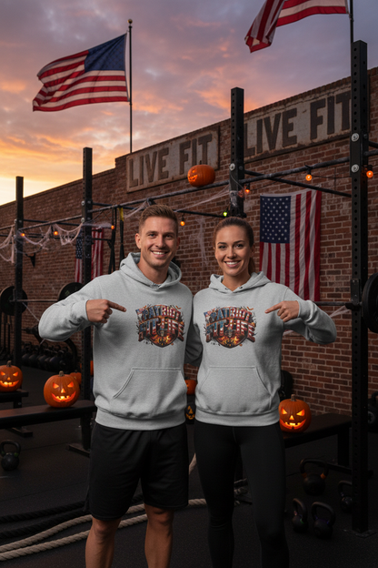 A man and a woman standing in a gym wearing matching light gray hoodies with a Halloween patriotic graphic print that reads 'Patriot Fit Life' with a collage of American flags. The background features a brick wall, American flags, weight racks, and Halloween decorations, including pumpkins.