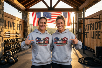 Two women standing in a rustic gym wearing matching PATRIOT FIT LIFE dictionary-style definition light gray hoodies with a patriotic American flag eagle graphic on the chest, pointing at the designs. The background features wooden walls, dumbbells, kettlebells, and American flags hanging in the distance.