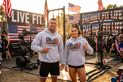 A man and a woman standing in an outdoor gym wearing matching PATRIOT FIT LIFE dictionary-style definition light gray hoodies with a patriotic American flag eagle with American flag-patterned wings graphic on the chest, pointing at the designs. The background features wooden walls, LIVE FIT and PATRIOT FIT LIFE signs, dumbbells, kettlebells, and American flags hanging in the distance.