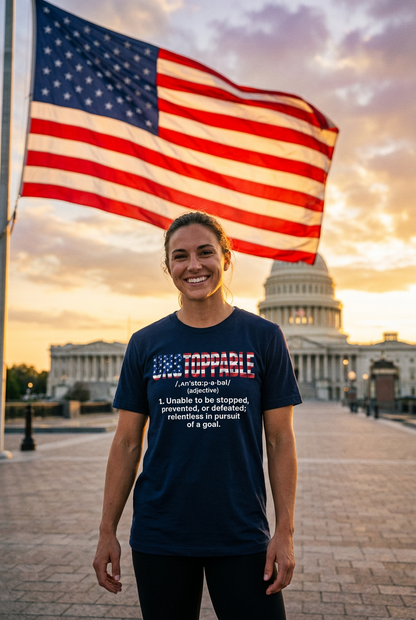A smiling woman stands outdoors wearing an UNSTOPPABLE navy blue t-shirt in front of the U.S. Capitol with an American flag flying above.