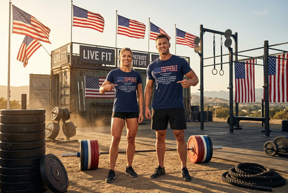 A man and a woman standing outdoors at a CrossFit-style gym, both wearing matching navy blue t-shirts with white text and logos. The background features American flags, a 'LIVE FIT' container, weight racks, and barbells.