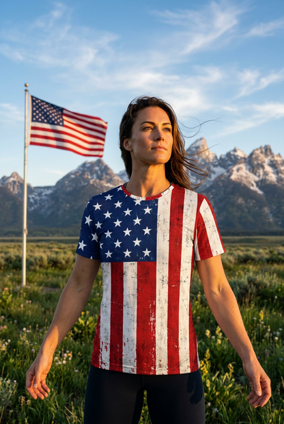 A woman stands outdoors in a grassy field with snow-capped mountains and a flag in the background, wearing a short-sleeve athletic shirt printed with a distressed American flag design.