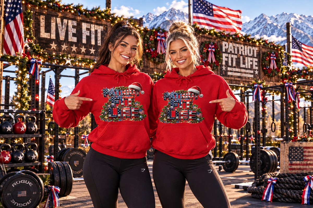 Two women stand in a decorated CrossFit gym wearing matching red hoodies with a festive American flag graphic on the front that reads PATRIOT FIT LIFE, pointing at the designs. The background features mountain scenery, 'LIVE FIT' and 'PATRIOT FIT LIFE' signs, and various fitness equipment.