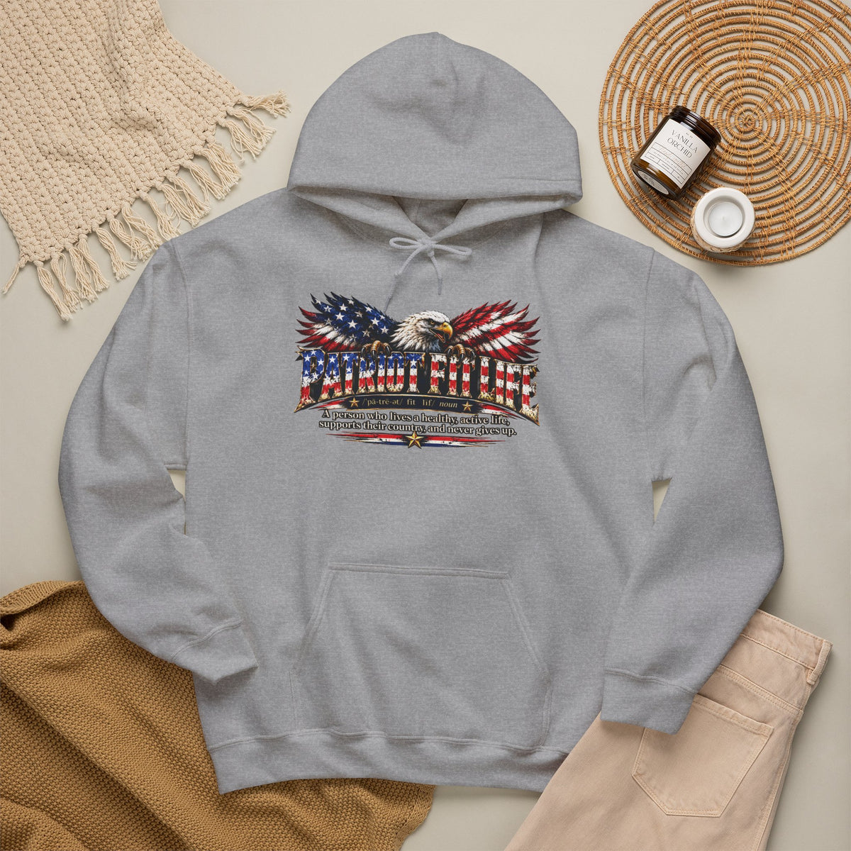 A light gray pullover hoodie laid flat on a beige surface, featuring a large patriotic graphic of an eagle with American flag-patterned wings and the text PATRIOT FIT LIFE with additional dictionary-style definition text below. 