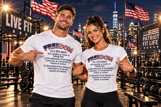 Two smiling people standing in a gym at night wearing matching white graphic T-shirts with a red, white, and blue 'FREEDOM' definition text design and pointing to the text on the shirts. The background shows a New York City skyline with flags and gym equipment.