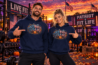 A man and a woman standing in an outside gym at sunset wearing matching navy blue hoodies with a large Halloween-themed patriotic PATRIOT FIT LIFE American flag pattern text graphic across the chest. The background includes workout equipment, American flags, banners reading 'LIVE FIT' and 'PATRIOT FIT LIFE,' and autumn decorations.