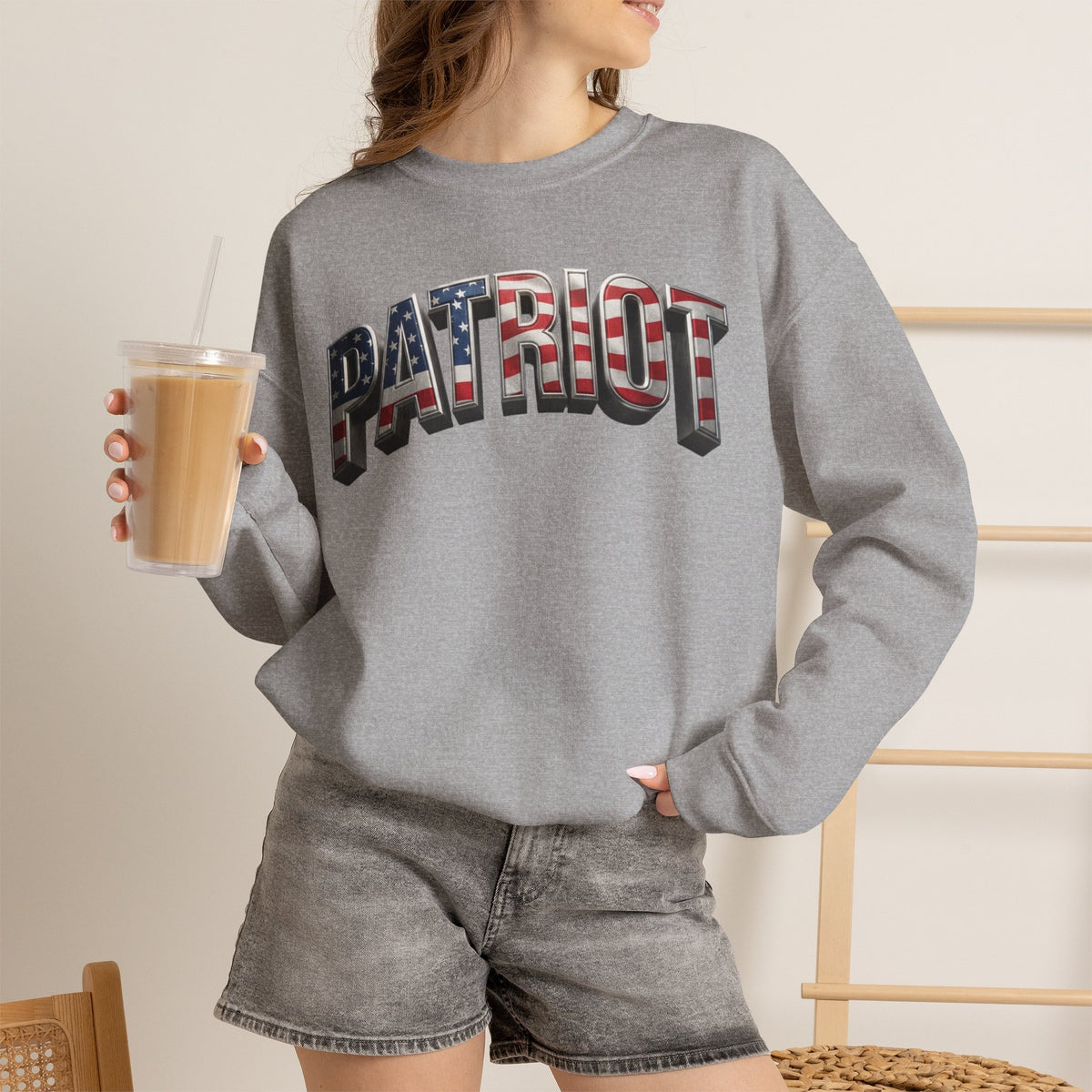 A woman wearing a heather gray crewneck sweatshirt with 'PATRIOT' printed across the chest in an American flag pattern, paired with gray denim shorts and holding an iced coffee.