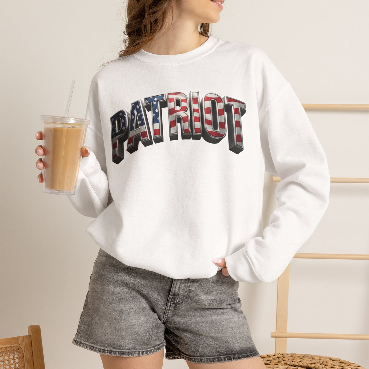 A woman wearing a white crewneck sweatshirt with a large 'PATRIOT' graphic across the chest filled with an American flag pattern, paired with gray denim shorts and holding an iced coffee.
