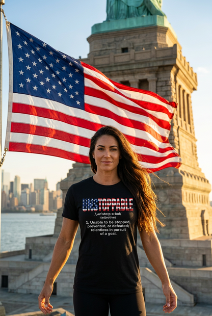 A woman stands in front of the Statue of Liberty wearing a black graphic T-shirt with a patriotic red, white, and blue design that reads 'UNSTOPPABLE' and a definition underneath. A large American flag is waving behind her, with the city skyline and statue visible in the background.