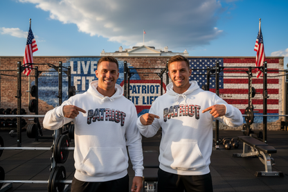 Two men standing in a gym, pointing at white hoodies with 'PATRIOT' printed across the chest. Behind them is a brick wall with a large American flag mural, weight racks, and two American flags in the sky.