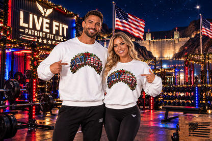 A man and a woman standing in a brightly lit gym at night wearing matching white crewneck sweatshirts with a colorful 'PATRIOT' graphic design across the chest. Behind them is the Hoover Dam, American flags, a large 'LIVE FIT and PATRIOT FIT LIFE' sign, weight equipment, and mountains under a starry sky.