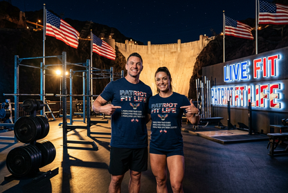 Two fitness coaches standing in front of the Hoover Dam at night wearing matching navy blue 'PATRIOT FIT LIFE' t-shirts and shorts, with American flags, gym equipment, and a neon sign in the background.