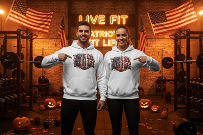 Two people standing in a gym wearing matching white hoodies with a Halloween patriotic graphic print on the front that reads "Patriot Fit Life" pointing at the chest design. The background features a brick wall, neon sign, American flags, weight racks, and jack-o'-lanterns.