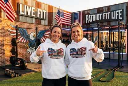 Two women standing in a gym wearing matching white hoodies with a patriotic PATRIOT FIT LIFE American flag eagle graphic on the front. The background features a brick wall with a painted bald eagle, American flags, and gym equipment under signs reading 'LIVE FIT' and 'PATRIOT FIT LIFE'.
