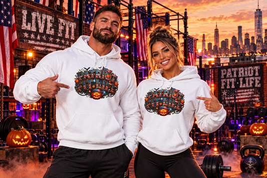 A man and a woman pose together in a gym setting at sunset, wearing matching white hoodies with a large patriotic Halloween-themed graphic on the chest with PATRIOT FIT LIFE text. The background features gym equipment, American flags, jack-o'-lanterns, and a city skyline at dusk.