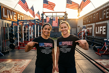 Two women standing in a gym wearing matching black t-shirts with a patriotic American flag graphic and the word 'UNSTOPPABLE' printed on the front. The background features weight equipment, American flags, and a warm sunset glow.