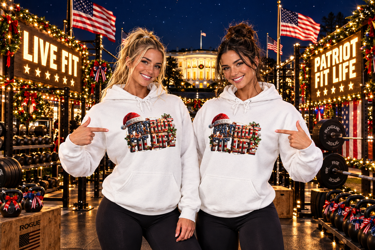 Two women wearing matching white graphic hoodies stand in a decorated gym at night, pointing to the holiday 'Patriot Fit Life' design printed on the front. The background features warm lights, American flags, and fitness equipment.