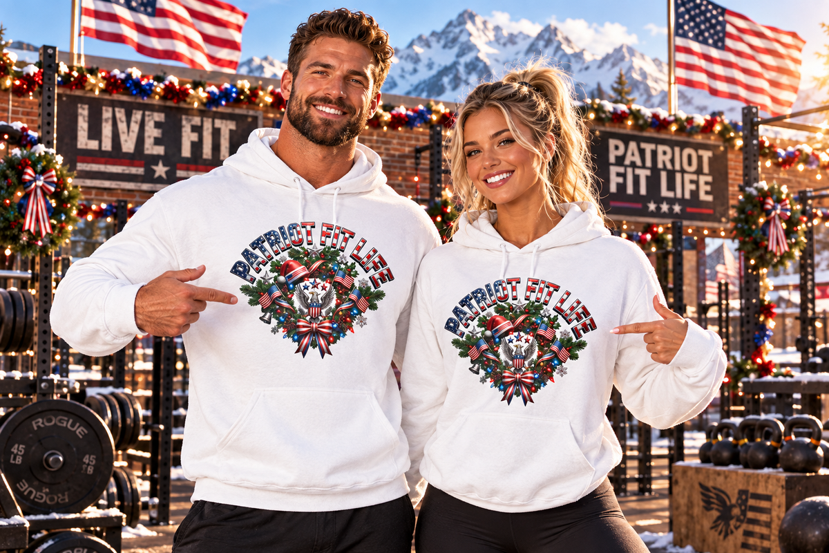 A man and a woman stand in a snowy outdoor fitness setting wearing matching white hoodies with a large patriotic festive graphic that reads 'PATRIOT FIT LIFE' and features an American flag, stars, and holiday wreath design. The background includes mountains, American flags, workout equipment, and branded signs.