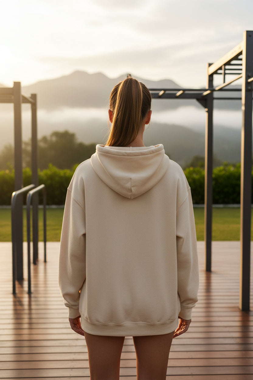 A woman wearing a beige hoodie standing on a wooden deck with mountains in the background.
