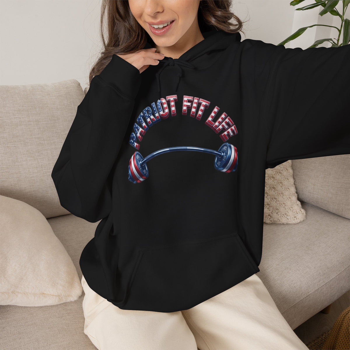 A woman wearing a black pullover hoodie with a patriotic-themed graphic that reads 'PATRIOT FIT LIFE' above a barbell with red, white, and blue flag-pattern details. She is sitting on a light beige couch in a home setting.