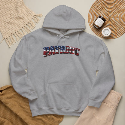 A heather gray pullover hoodie laid flat with a front kangaroo pocket and a large arched 'PATRIOT' graphic across the chest in an American flag pattern. The hoodie is styled in a flat lay with a woven placemat, small jar, knit blanket, tan sweater, and light pants on a light beige background.