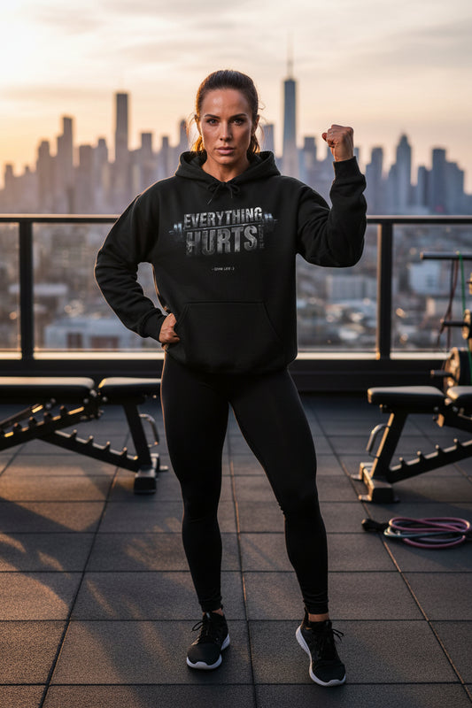 A woman wearing a black hoodie with text that says Everything Hurts! - GYM LIFE :) standing on a rooftop with a city skyline in the background.